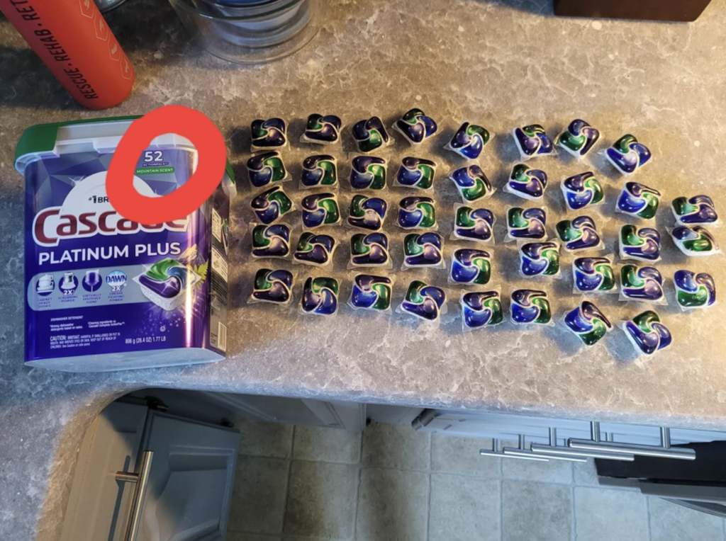 A package of 52 Cascade Platinum Plus dishwasher pods is open on a countertop, and approximately 42 individual pods are placed in a grid-like arrangement next to it. The pods are green, blue, and white in color.