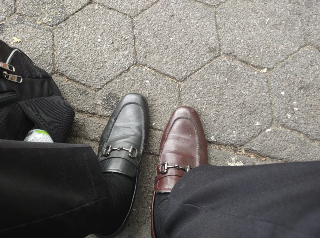 A person is wearing mismatched dress shoes—one black and one brown—while standing on a hexagonal tile pavement. Part of a black bag with zippers is visible on the left side.