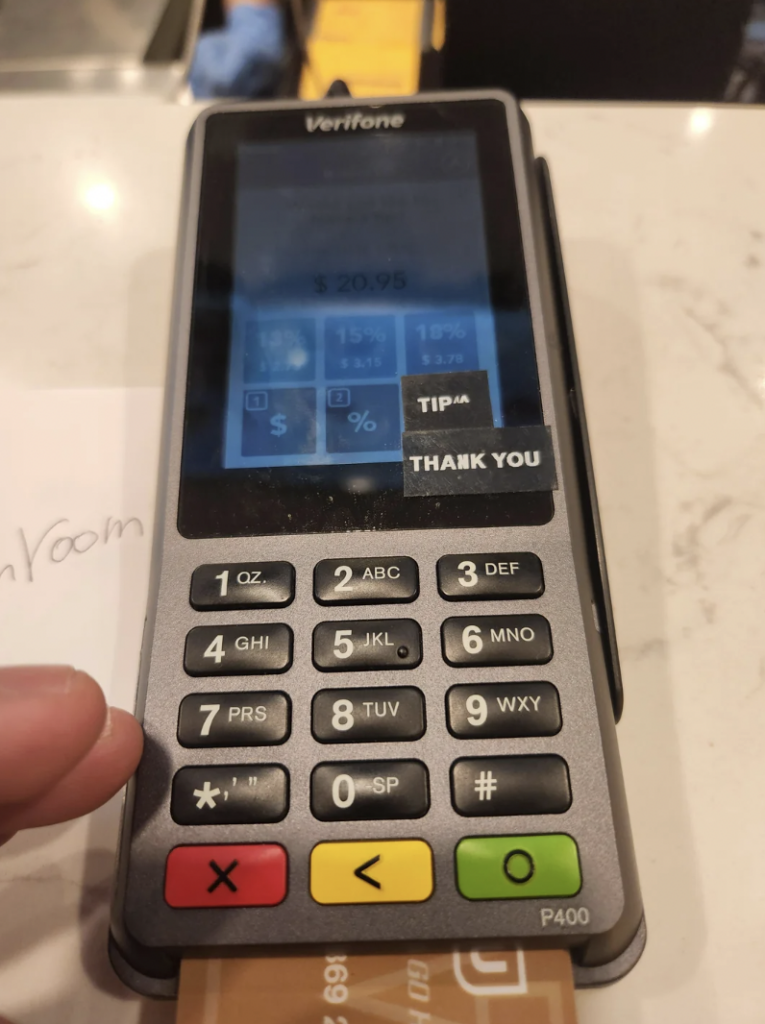 A Verifone payment terminal displays a total of $20.95. The screen shows tip options and a "Thank You" message. The terminal's keypad includes alphanumeric keys, function buttons, and a colored circle for confirmation. A hand is inserting a card at the bottom.