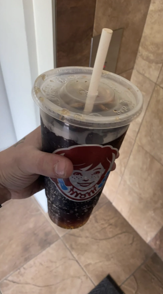 A hand holds a large plastic cup filled with a dark fizzy drink and a white straw. The cup features the Wendy's logo with the face of the character wearing pigtails. The background shows a tiled floor and part of a wall.