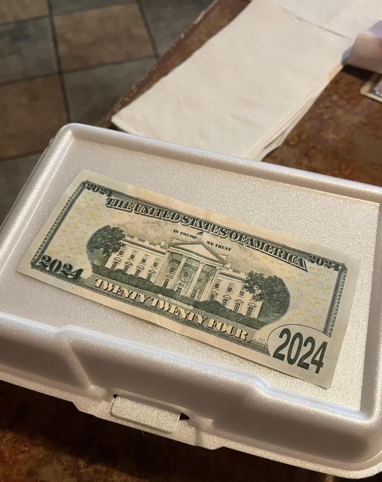 A United States 20-dollar bill featuring "2024" in large numerals on a white takeout container with a slightly textured surface. The bill lies flat on the container, which is on a brown countertop with napkins in the background.