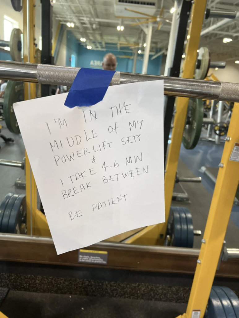 A handwritten sign taped to a barbell in a gym reads: "I'M IN THE MIDDLE OF MY POWERLIFT SETS. I TAKE 4-6 MIN BREAK BETWEEN. BE PATIENT." The sign is affixed with blue tape, and gym equipment is visible in the background.