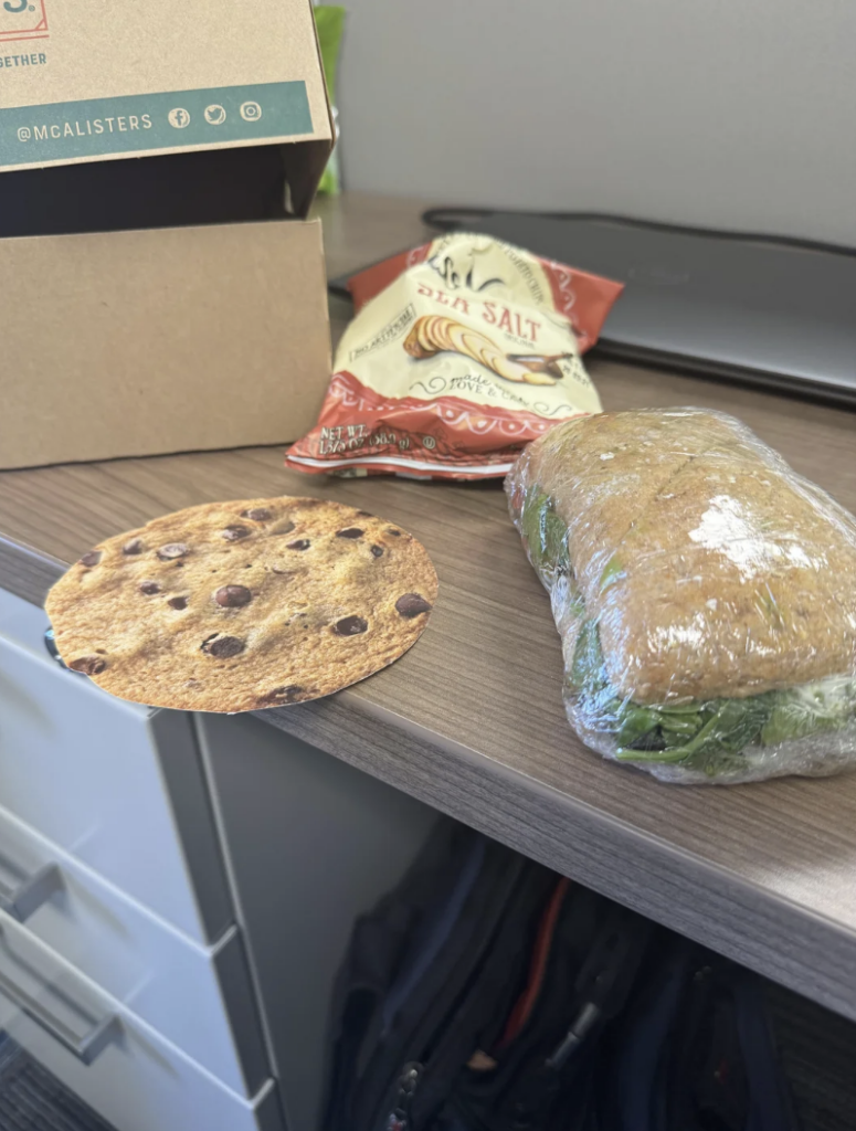 A desk with a chocolate chip cookie, a bag of kettle-cooked sea salt chips, and a sandwich wrapped in plastic. A cardboard box and a closed laptop are in the background.