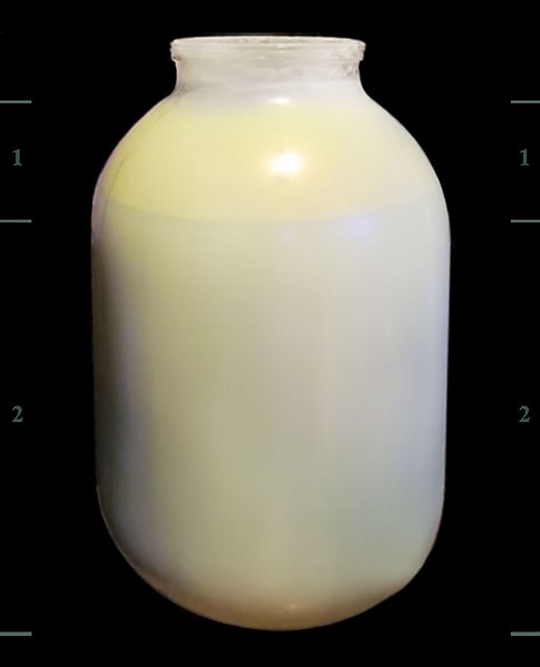 A large, opaque, egg-shaped container filled with a light yellowish liquid is shown against a black background. The container has a cylindrical neck and no visible markings. Small, numbered vertical scales (labeled 1, 2) are faintly visible on either side.