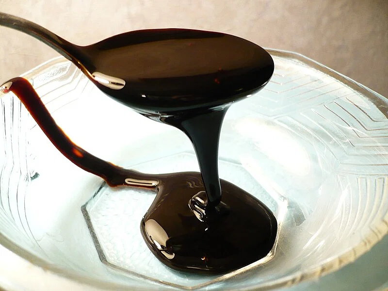 Close-up of a spoon drizzling thick, dark molasses into a glass bowl. The molasses forms a smooth, glossy ribbon as it is poured from the spoon, and the bowl features a geometric pattern. The background is softly blurred, keeping the focus on the molasses.