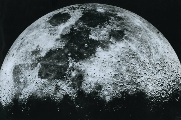 Black and white high-resolution photograph of the Moon, showing rugged surface details including craters and varied surface textures. The image appears to be taken from close proximity, capturing the intricate lunar landscape.