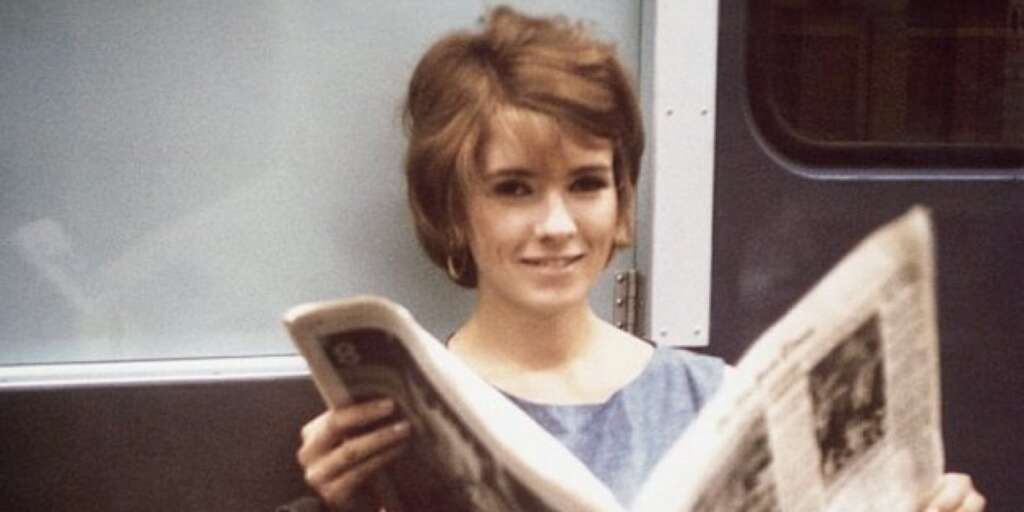 A young woman with short brown hair is sitting and smiling while holding and reading a newspaper. She is wearing a light blue top and is seated in what appears to be a train or subway with a window behind her.