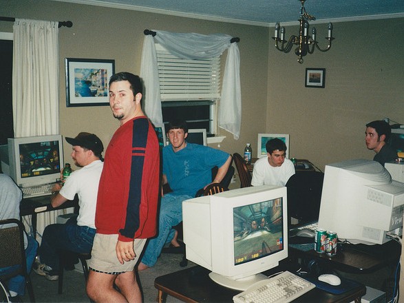 Several people in a room participate in a LAN party, playing computer games on desktop computers. One person stands in the foreground, while others sit focused on their screens. The room features typical home decor and has a relaxed atmosphere.