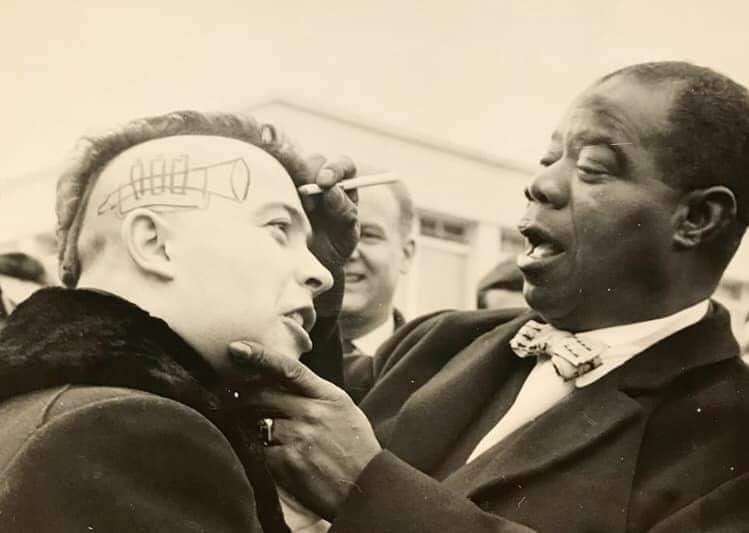 A black and white photo of two men. The man on the right, dressed in a suit with a bow tie, appears to be drawing on the shaved head of the man on the left with a writing instrument. The man on the left leans forward, smiling. People are in the background.