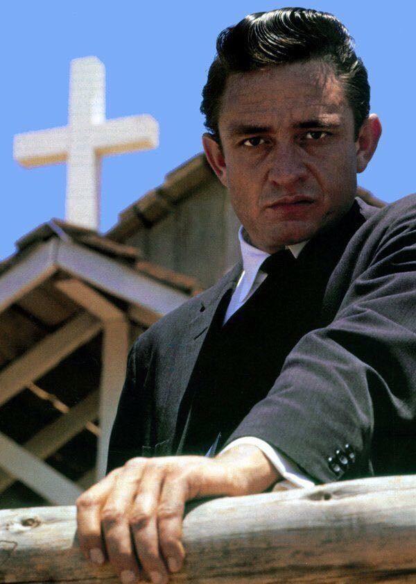 A man in a black suit and white shirt stands outside, leaning on a wooden railing with a serious expression. A cross and part of a church with a steep roof are visible in the background against a clear blue sky.