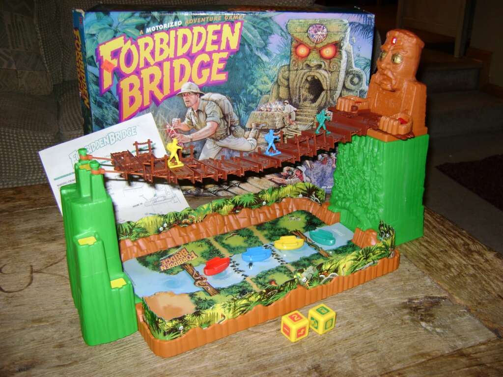 A board game called “Forbidden Bridge” is set up on a table. The game features a bridge suspended between two cliffs with colorful player pieces and dice. The box in the background depicts an adventurer crossing a bridge, guarded by a giant stone figure.