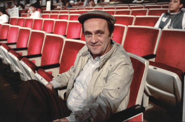 A man wearing a flat cap and a light-colored jacket sits in a row of red and white seats in a large indoor venue. He has a relaxed expression and one arm resting on the back of an adjacent seat. Other people are seated in the background.