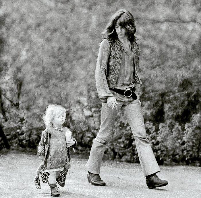 A man with long hair wearing a patterned vest and bell-bottom pants walks beside a young child with curly hair, who is holding a bottle. The scene is outdoors, with trees and bushes in the background. The image is in black and white.