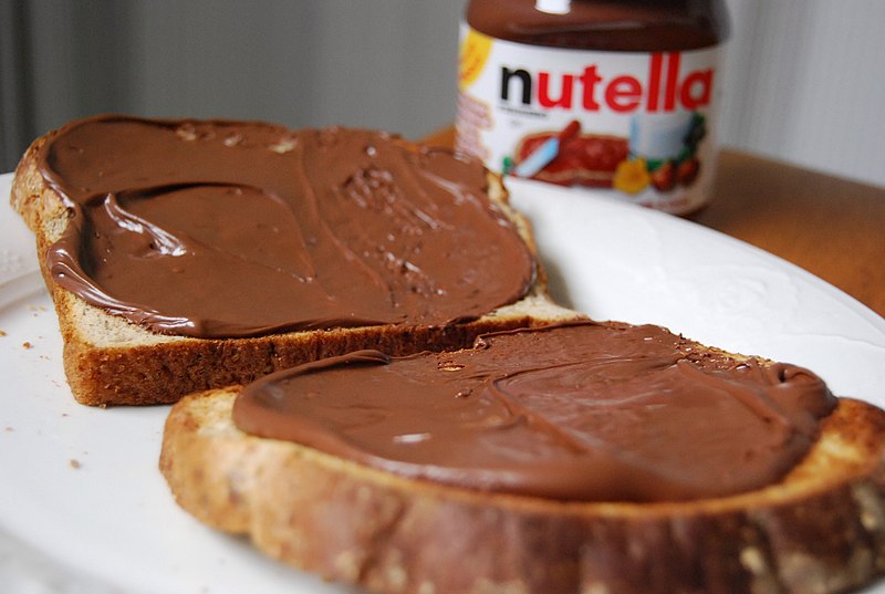 Two slices of bread are spread with Nutella hazelnut spread, placed on a white plate. A jar of Nutella is slightly blurred in the background.