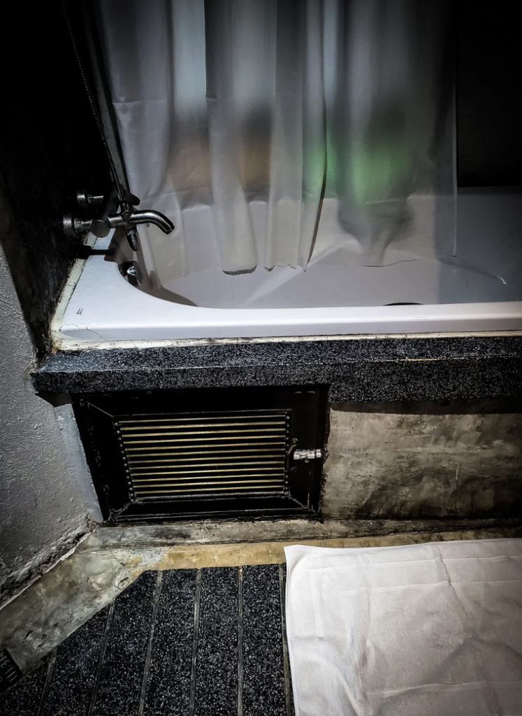 A bathroom with a dark granite bathtub surround, a white shower curtain partially closed, and a bath mat on the tile floor. The focus is on the tarnished metal vent positioned below the tub, set into a concrete wall.