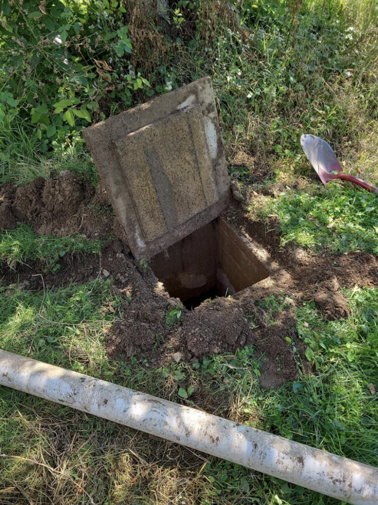 A square hole in the ground with a concrete cover leaning to the side, surrounded by dirt and grass. A shovel is stuck upright in the soil beside the hole, and a long metal pipe lies on the grass nearby. Trees and foliage are visible in the background.