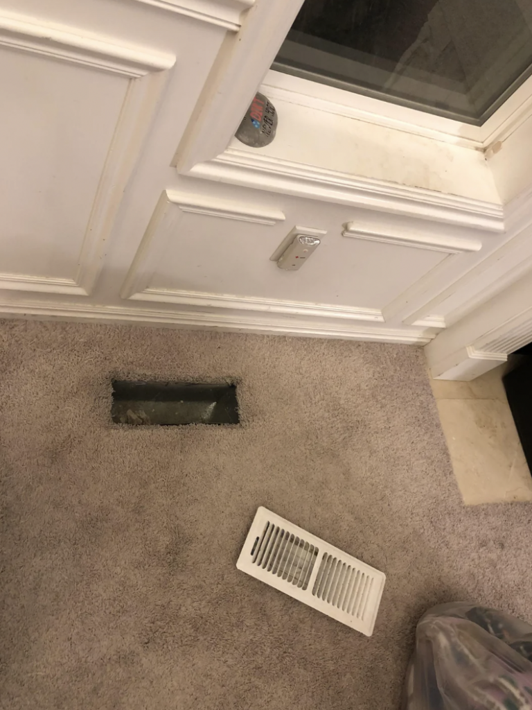 A partially installed air vent is visible on a carpeted floor with a white vent cover positioned next to it. The white baseboard in the background has detailed molding and there is a window or door above the baseboard with a latch.