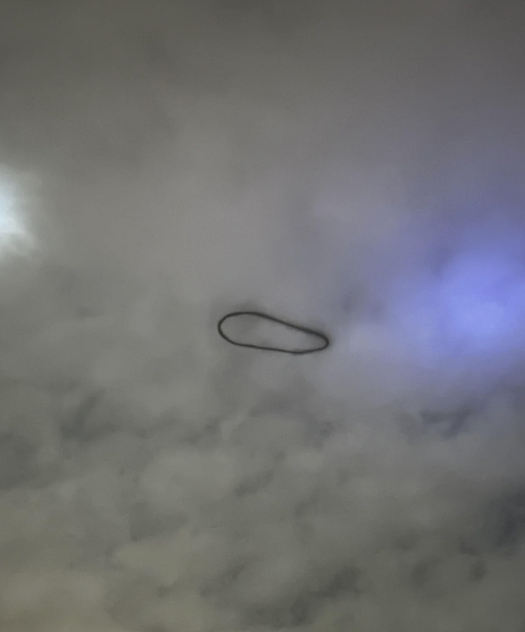 A dark, elongated oval shape is floating in a cloudy sky, illuminated faintly by a blue and white light in the background. The overall atmosphere is mysterious and ethereal.