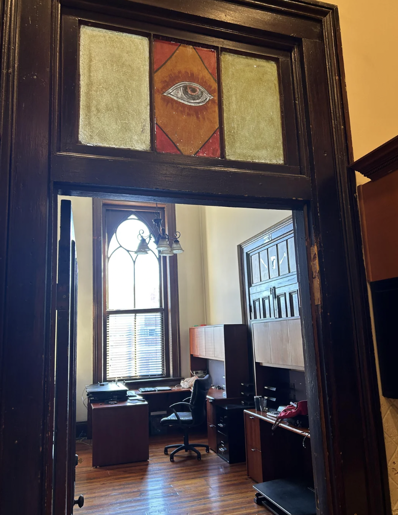 A view through a doorway into an office with wooden flooring and furniture. Above the doorway is a stained glass window featuring an eye design. Inside the office is a desk, chairs, shelves, and a large window with blinds.