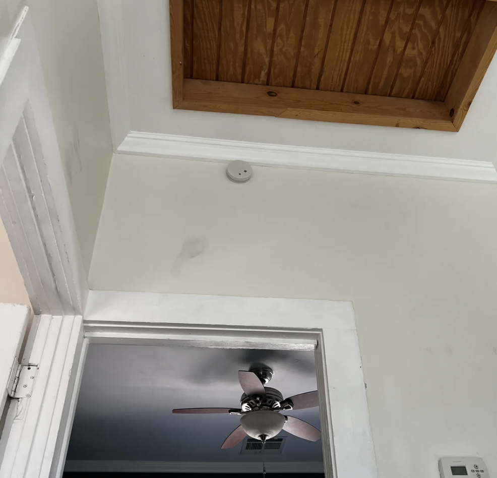 This image shows a section of a room with a ceiling fan and light visible through an open door. The white wall has a wooden ceiling panel and a small round smoke detector or alarm mounted near the ceiling. There is also a thermostat on the wall.