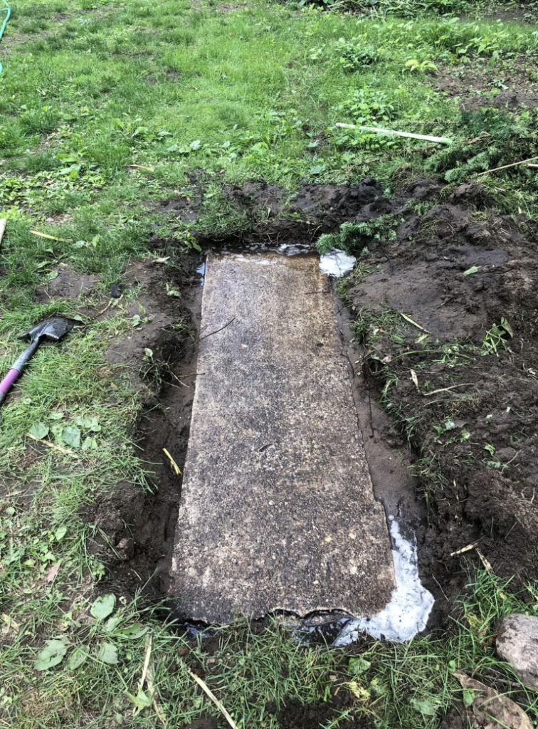 A large, rectangular concrete slab is partially embedded in the ground with dirt and grass around it. A shovel with a purple handle rests nearby, suggesting ongoing excavation work. The surrounding area is green with patches of grass and plants.