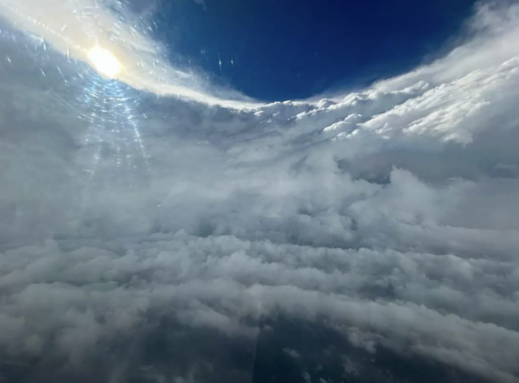 A spectacular view from an airplane window shows a partly cloudy sky with the sun breaking through, casting rays of light across the clouds below. The horizon line is visible, separating the blue sky above from the dense, cotton-like clouds below.