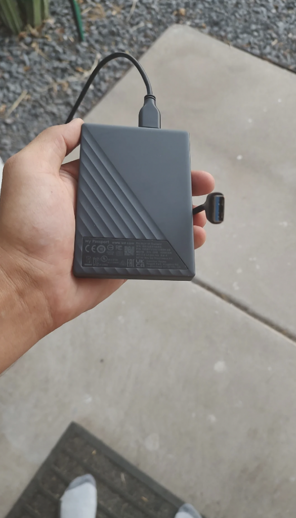 A hand is holding a black portable external hard drive connected via USB cable. The drive has a textured diagonal design and visible labels with technical specifications on the bottom half. The background features a concrete surface and gravel.