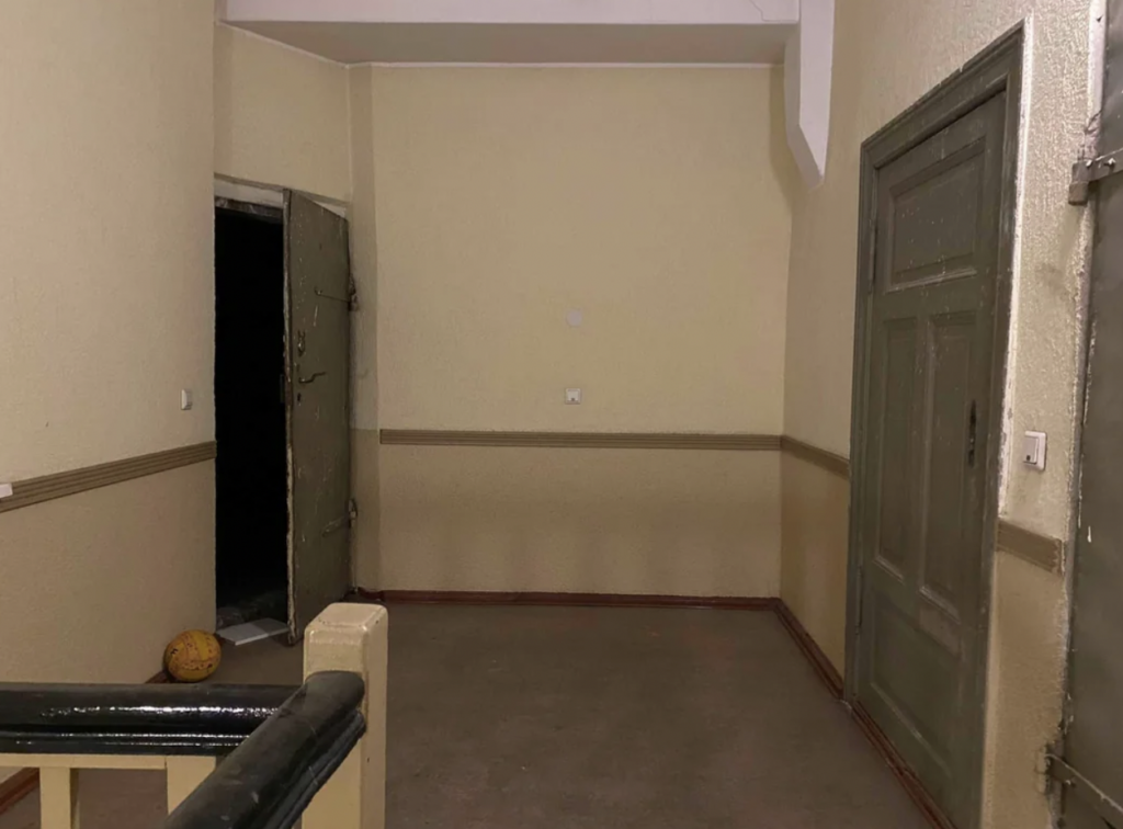 An hallway with cream-colored walls and a beige flooring. To the left, a dark doorway stands open with a pumpkin-like object on the floor near it. On the right, a closed door is visible. A stair railing can be seen in the foreground.