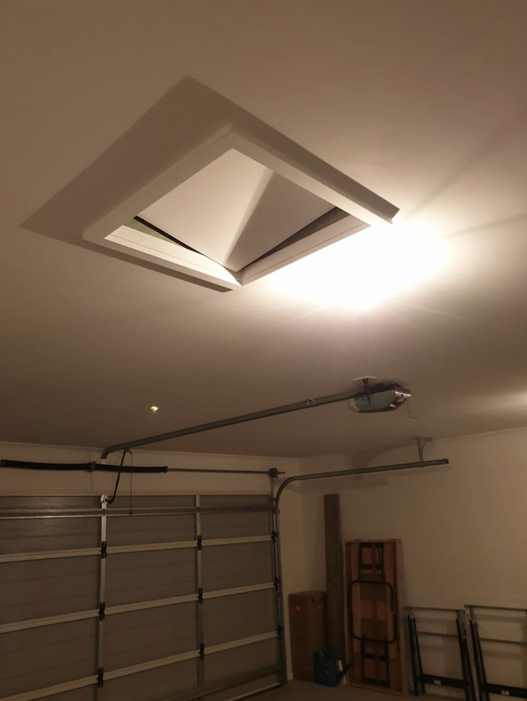 A ceiling access panel partially open in a garage with overhead lighting illuminating the space. The garage door in the background is closed. Various items are partially visible around the garage, including folded tables and storage items.