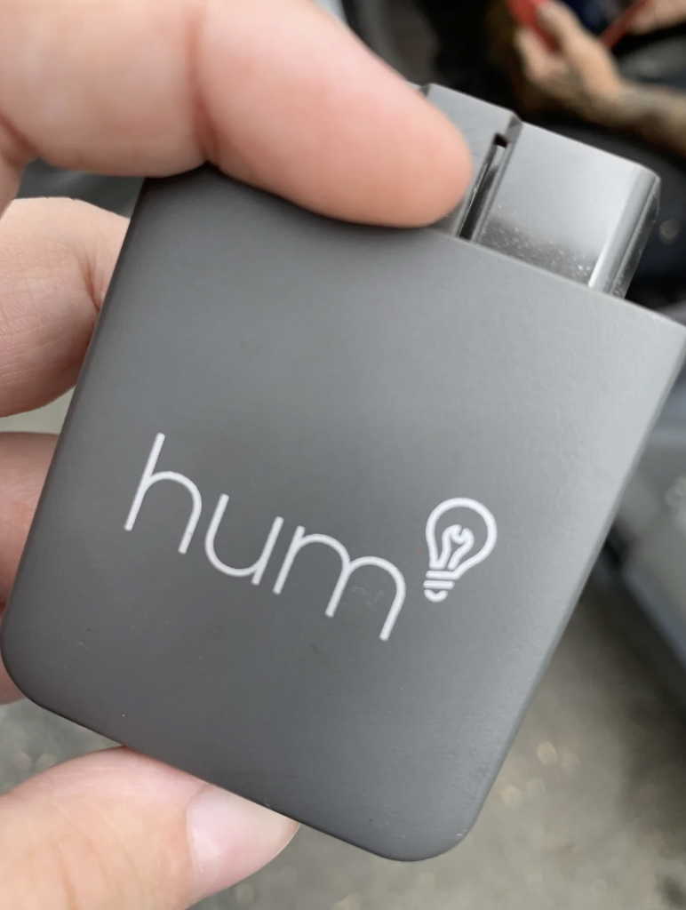 A hand holds a small, grey electronic device with the word "hum" and an icon of a light bulb printed on it.