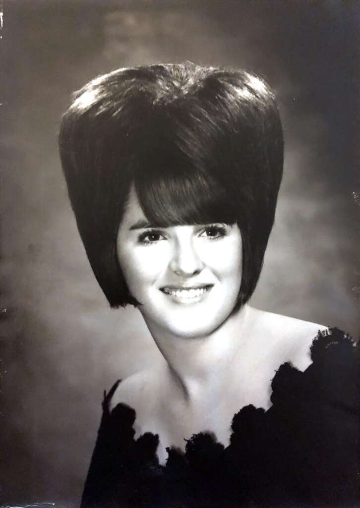 A black and white portrait of a woman with a large, voluminous hairstyle. She has a fringe (bangs) and is smiling. She is wearing an off-the-shoulder top with floral details. The background is plain and blurred.