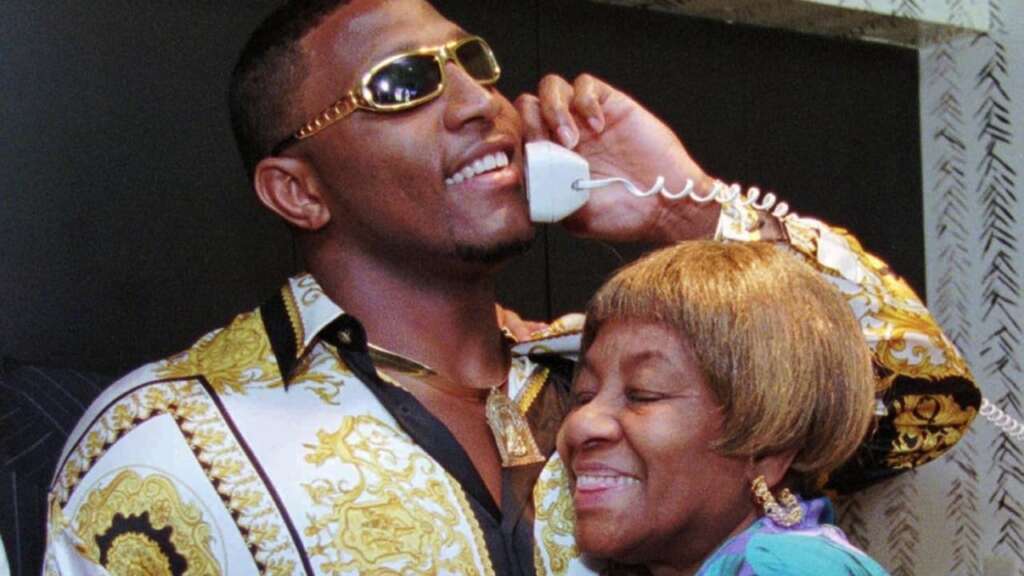 A man wearing sunglasses and a patterned shirt with gold designs is talking on a corded phone while smiling. An older woman with short hair and a striped shirt is hugging him from the side, smiling warmly. They appear to be happy and enjoying the moment.