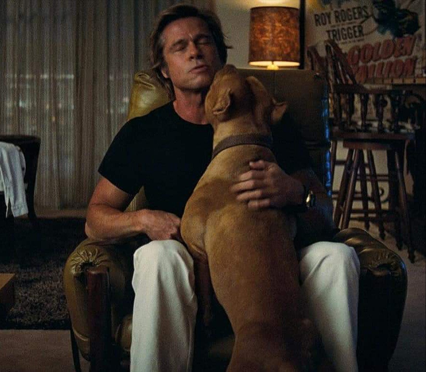 A man with blonde hair, wearing a black T-shirt and white pants, sits in an armchair with his eyes closed, while a large brown dog stands with its front paws on his lap, nuzzling his face. The background shows a lamp and a vintage poster on the wall.