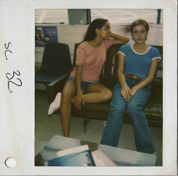 Two young women are sitting on a couch. The woman on the left, wearing a pink shirt and denim shorts, is leaning towards the woman on the right and whispering in her ear. The woman on the right, wearing a blue shirt and jeans, sits with her hands in her lap, looking forward.