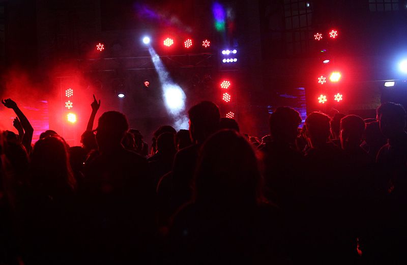 A crowd of people is silhouetted against a series of bright red and white lights at a concert or music event. The atmosphere is energetic with people raising their hands and the stage lights cutting through smoke or fog.