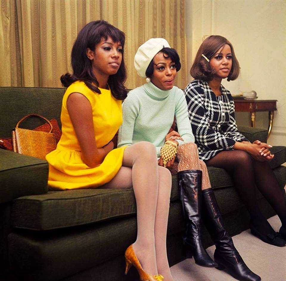 Three women are seated on a green sofa in a room with a curtained window. The woman on the left wears a yellow dress, the middle one a light blue outfit with a white beret, and the right one a black and white checkered dress. They all have stylish hairstyles.