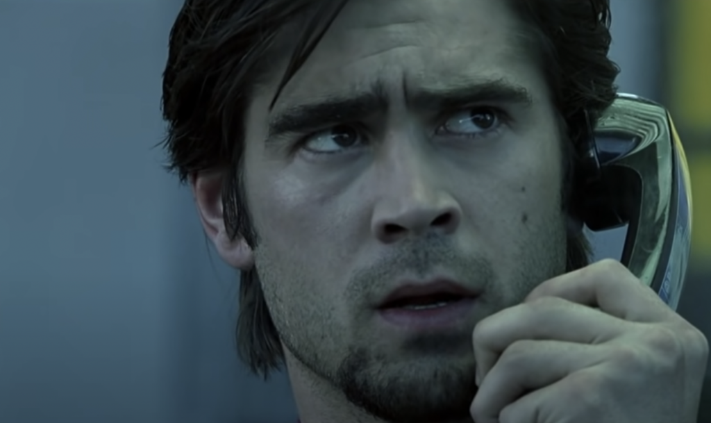 A man with dark hair and a slight beard holds a phone receiver to his ear, looking seriously concerned. He has a furrowed brow and slightly parted lips, as though he's speaking or listening intently. The background is blurred and has a cool-toned color scheme.