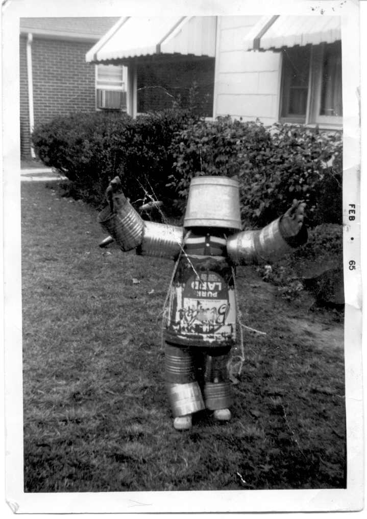 A black-and-white photo from February 1965 shows a child in a homemade robot costume. The outfit consists of a bucket helmet, cylindrical arms, and body made from tin cans and other metal parts. The child stands with arms raised in front of a house with a brick wall and bushes.