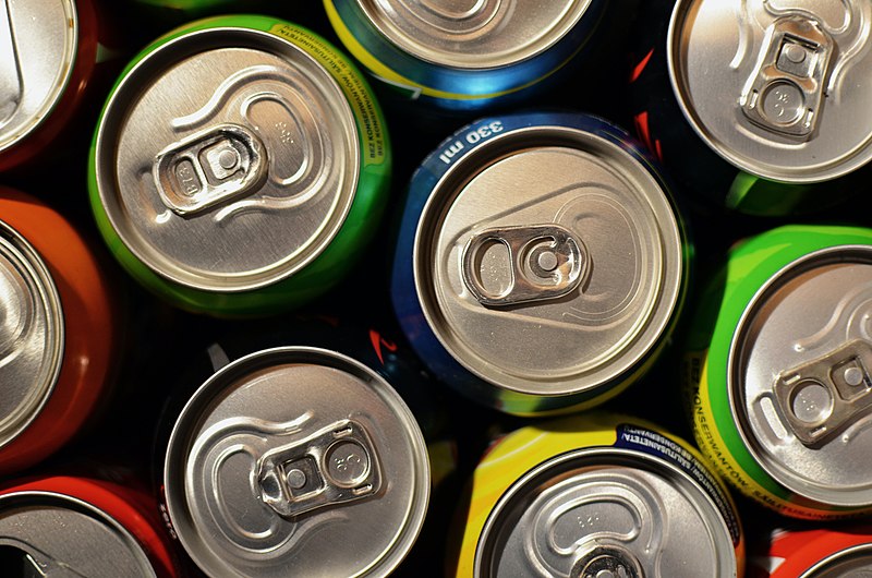 A close-up shot of the tops of various soda cans arranged in a tight group. The cans have different colors, suggesting different flavors and brands. Each can is equipped with a pull-tab for opening. The metallic surfaces reflect light, highlighting their contours.