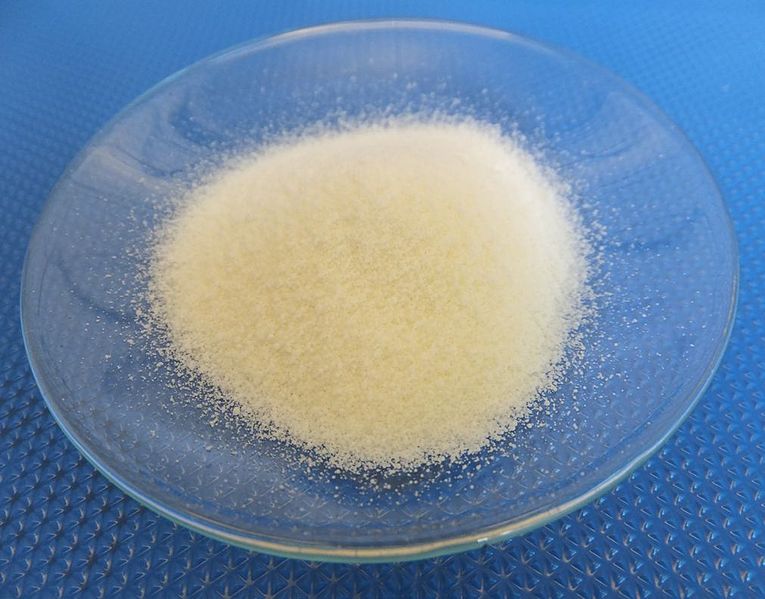 A small mound of white powder is displayed on a clear, round glass dish. The dish is placed on a textured blue surface. The fine granules are spread evenly across the center of the dish.