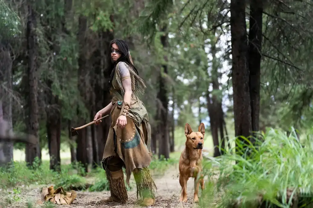 A person dressed in rustic, forest attire holds an axe and stands alertly in a dense forest, looking back over their shoulder. They are accompanied by a tan dog standing attentively beside them on the forest path. Tall trees and greenery surround them.