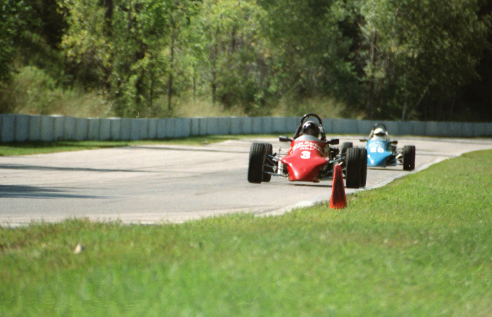 Two race cars approach a corner on a racetrack lined with greenery and safety barriers. The car in front is red with the number 3, followed by a blue car with the number 60. A cone is positioned on the edge of the track.