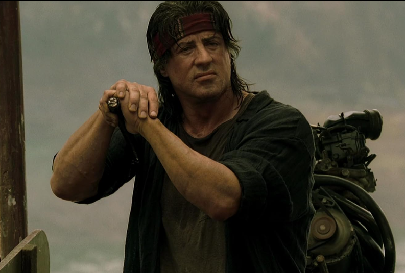 A rugged man with long, dark hair and a red headband stands outdoors, gripping a weapon. He is wearing a black shirt with the sleeves rolled up, and a tactical backpack is visible behind him. The background appears to be a misty, mountainous landscape.