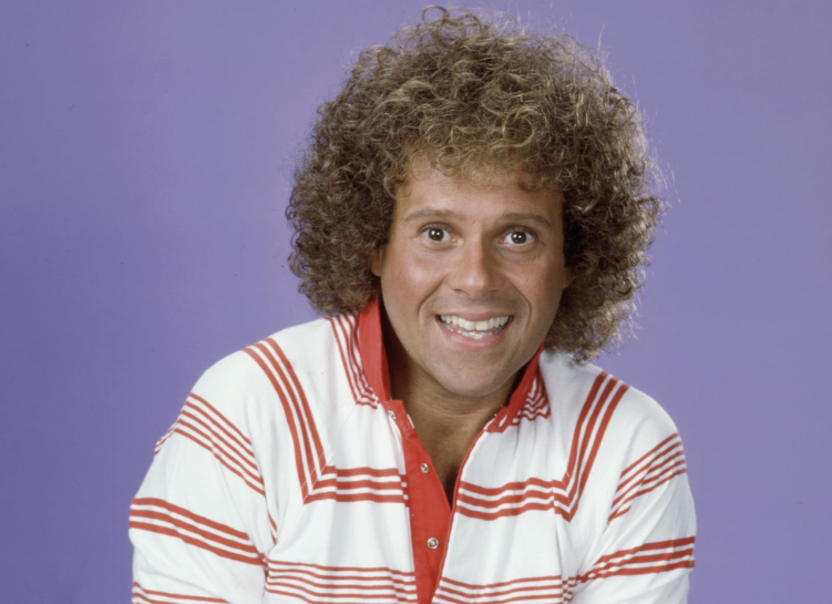 A man with curly hair, smiling broadly, wearing a white polo shirt with red horizontal stripes and a red collar, stands against a plain purple background.