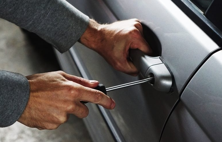 Close-up of a person attempting to break into a car using a screwdriver to manipulate the car's door handle. The individual is wearing a grey, long-sleeved shirt and the car is silver.