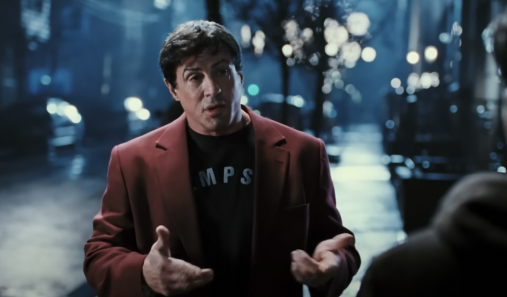 A man with dark hair, wearing a red blazer over a black shirt, stands outdoors at night in an urban setting. He is gesturing with his hands while facing the camera. The background is dimly lit with streetlights and trees, creating a moody atmosphere.