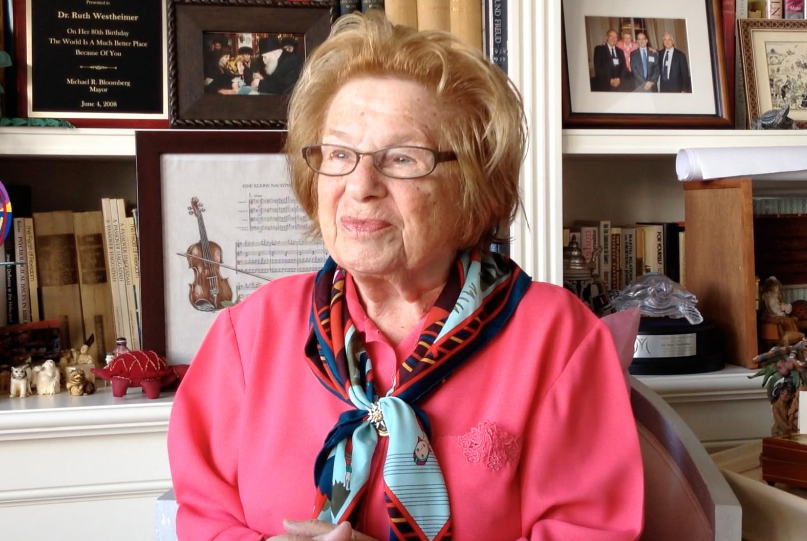 An elderly woman with short, light hair and glasses, wearing a bright pink sweater and a colorful scarf, is sitting in front of a bookcase filled with books, framed pictures, and small decorative items. She is smiling and looking slightly to the side.