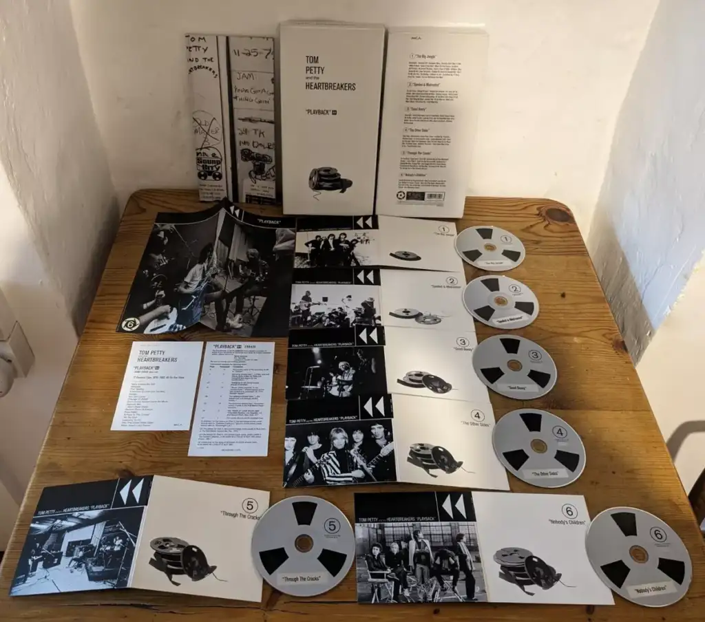 A collection of Tom Petty and the Heartbreakers memorabilia displayed on a wooden table. Items include vinyl records, CDs, booklets, posters, a setlist, and other printed materials. The overall color scheme is predominantly black and white.