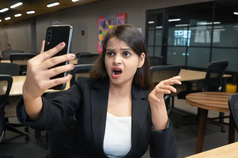 A woman with long brown hair, wearing a black blazer and white top, is sitting in an office and looking at her phone with a shocked and upset expression. Her mouth is open, and her left hand is raised with fingers spread out.