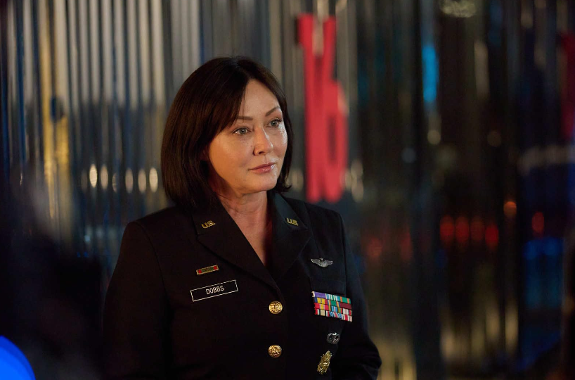 A person in a military uniform with numerous ribbons and medals stands in front of a corrugated metal background. The individual has straight brown hair and is looking to the side, wearing a name tag that says "Oberis." The surroundings appear to be dimly lit.